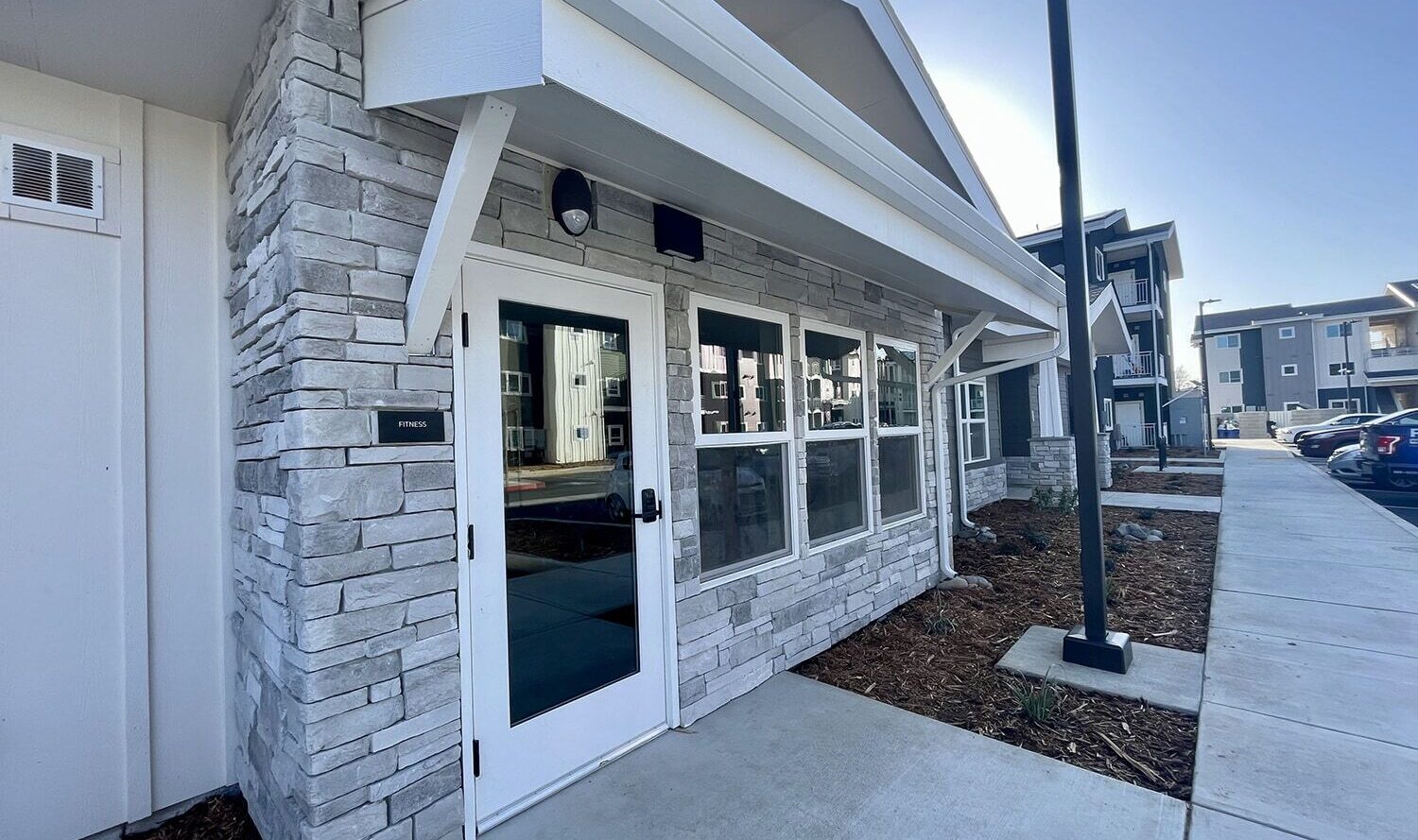 Entrance to a modern building with stone walls and multiple windows. A small awning covers the doorway, and a paved walkway leads to the entrance. Nearby are neatly landscaped areas and adjacent buildings with similar architecture.