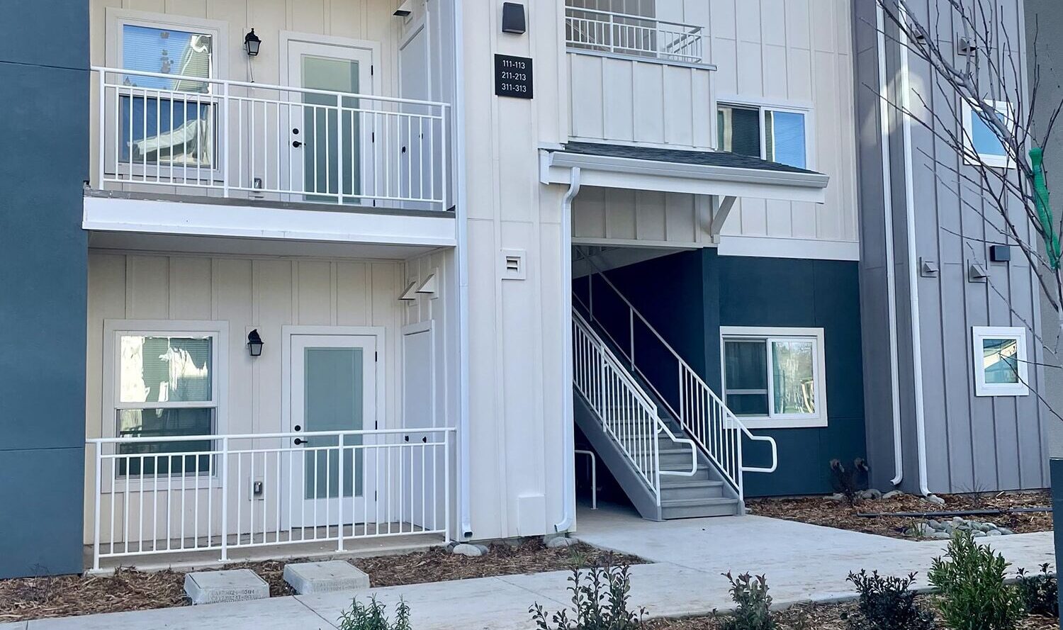 A modern three-story apartment building with white balconies and a combination of blue and white exterior walls. A small landscaped area with shrubs and stones is in the foreground, and the sky is clear and blue.