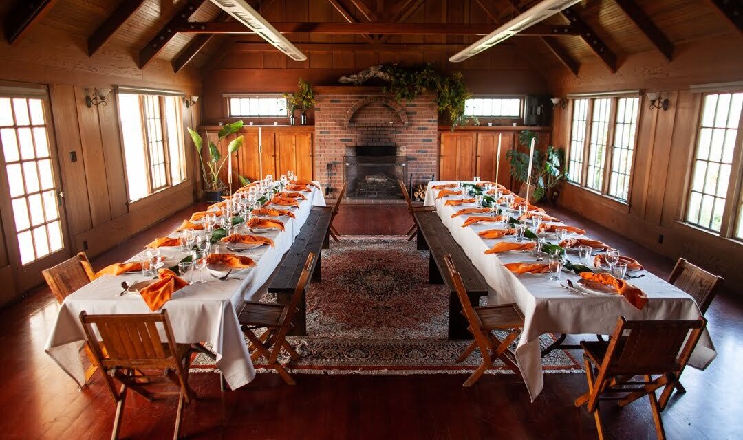 A cozy dining room with two long tables arranged in a U-shape, set with white tablecloths and vibrant orange napkins. Wooden chairs surround the tables, and a brick fireplace is centered on the back wall, surrounded by rustic wooden beams.