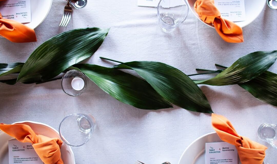 A table setting with white plates, orange napkins, and green leaves as a centerpiece. Glasses and cutlery are arranged neatly, and menu cards are visible on the plates.