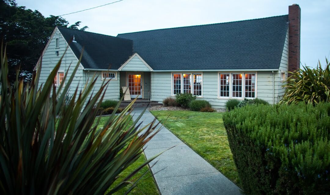A cozy, gray cottage with lit windows on a cloudy evening. A pathway leads through a neatly trimmed lawn, flanked by bushes and tall grasses. A tall tree is visible in the background, with soft ambient lighting adding warmth.