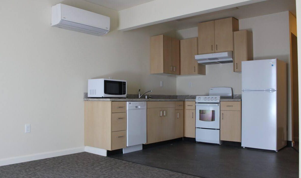 A small kitchen with wooden cabinets, a white refrigerator, microwave, stove, and dishwasher. Air conditioning unit mounted on the wall and a gray carpeted floor in the foreground. Walls are painted light beige.