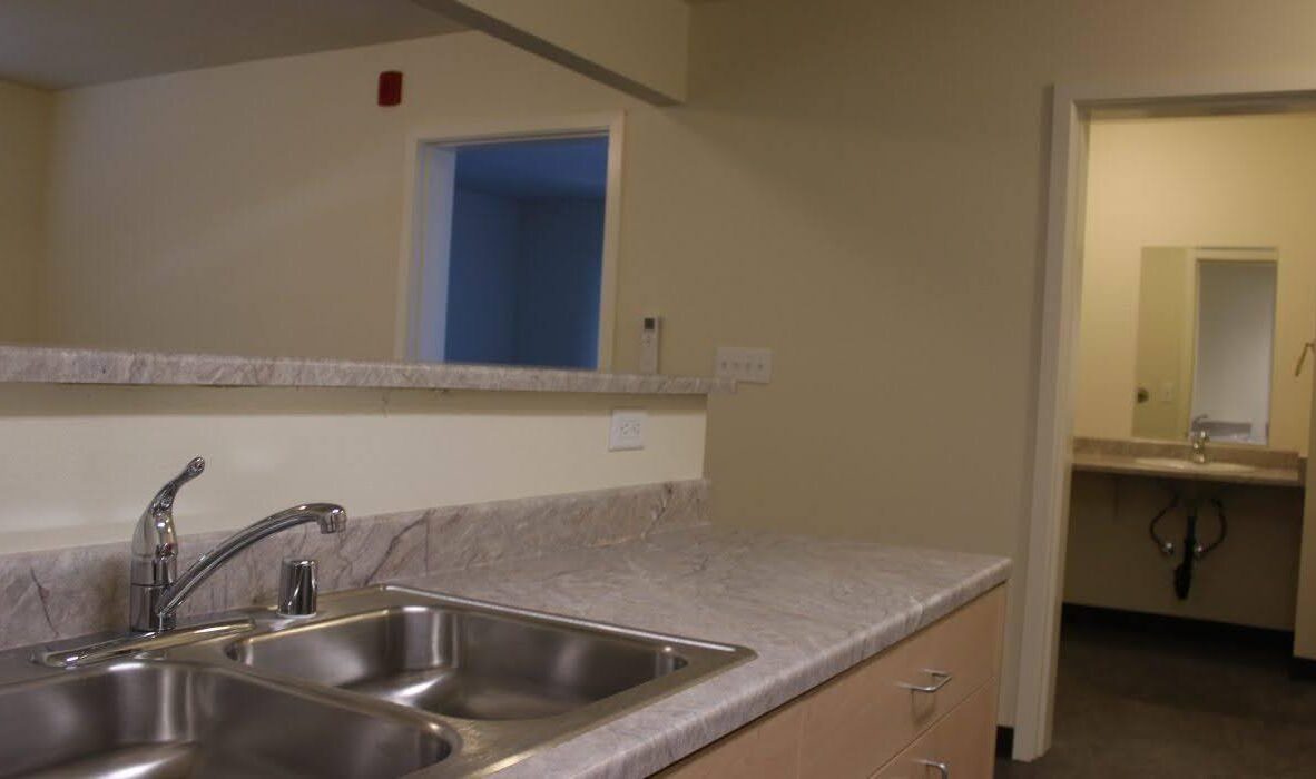 A modern kitchen featuring a dual sink with chrome faucet on a marble counter. Behind the counter is an open area leading to a room, with a view of a sink and mirror in another small room. The walls are painted a neutral color.