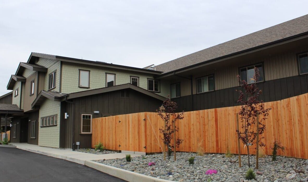 A modern two-story building with a mix of green and dark wood siding, next to a wooden fence. The foreground features a landscaped area with rocks and small plants. The sky is overcast.