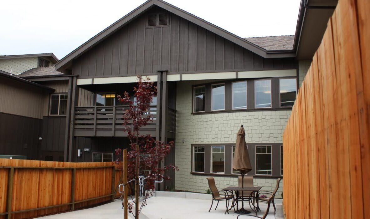 A modern two-story house features a wooden exterior with a covered balcony. The backyard includes a wooden fence, a patio table with chairs, and a closed umbrella. Young trees are planted around the paved patio area.