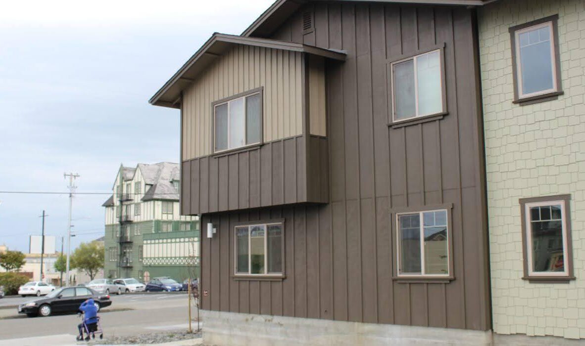 A two-story building with brown and cream siding stands on a street corner. A person in a blue jacket pushes a stroller on the sidewalk. Multiple buildings are visible in the background under an overcast sky.