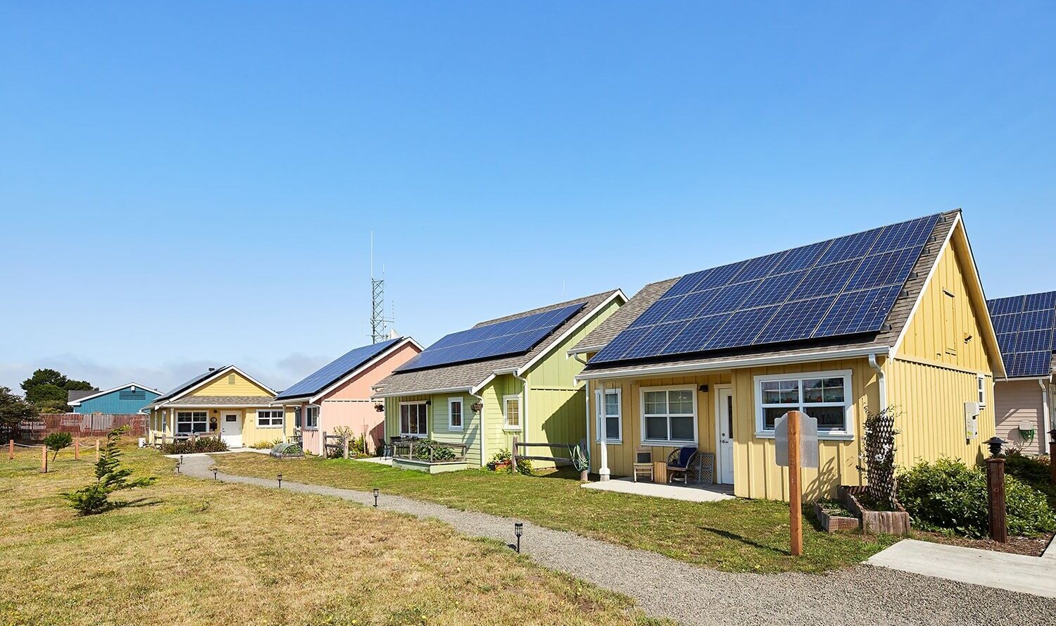 A row of colorful houses with solar panels on the roofs, set against a clear blue sky. The houses are painted yellow, green, pink, and blue, with a grassy area and pathway in front.