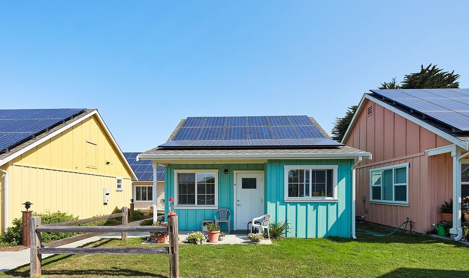 A small turquoise house with solar panels on the roof is flanked by a yellow house and a beige house, both also with solar panels. There is a white door and two windows at the front. The houses are set under a clear blue sky.