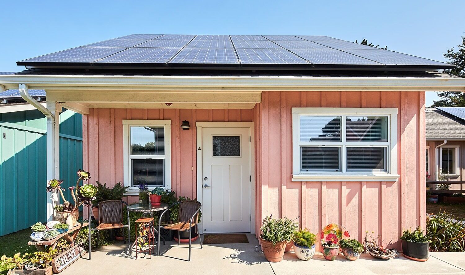 A small, pink house with solar panels on the roof. There’s a white door and a window with blinds. Several potted plants and a few chairs with a small table are arranged on the concrete patio. A Welcome sign is placed near the entrance.