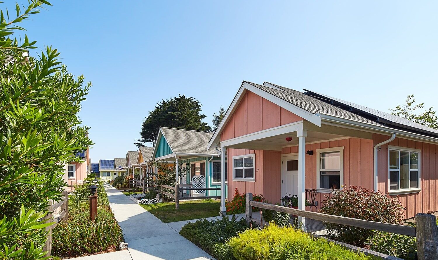 A row of small, colorful houses with front yards along a sidewalk. The sky is clear, and the plants and shrubs are well-maintained. One house has solar panels on the roof. The scene feels calm and inviting.