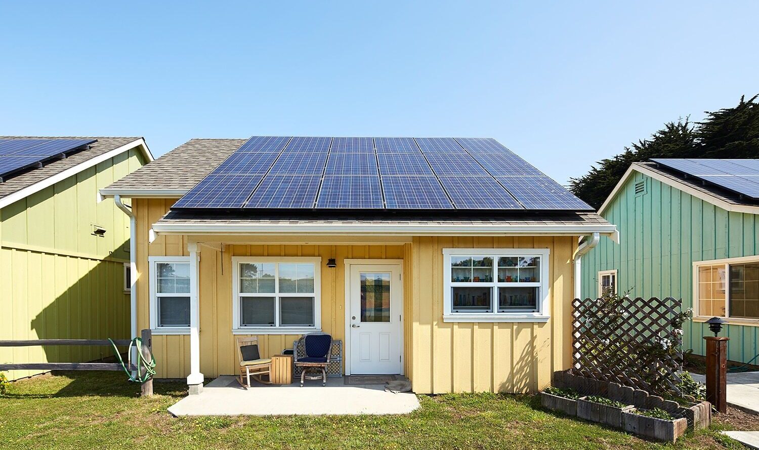 A small yellow house with solar panels on the roof. The front has a white door, two windows, and a patio with chairs. The lawn is green, and the neighboring house is light green. A clear blue sky is in the background.