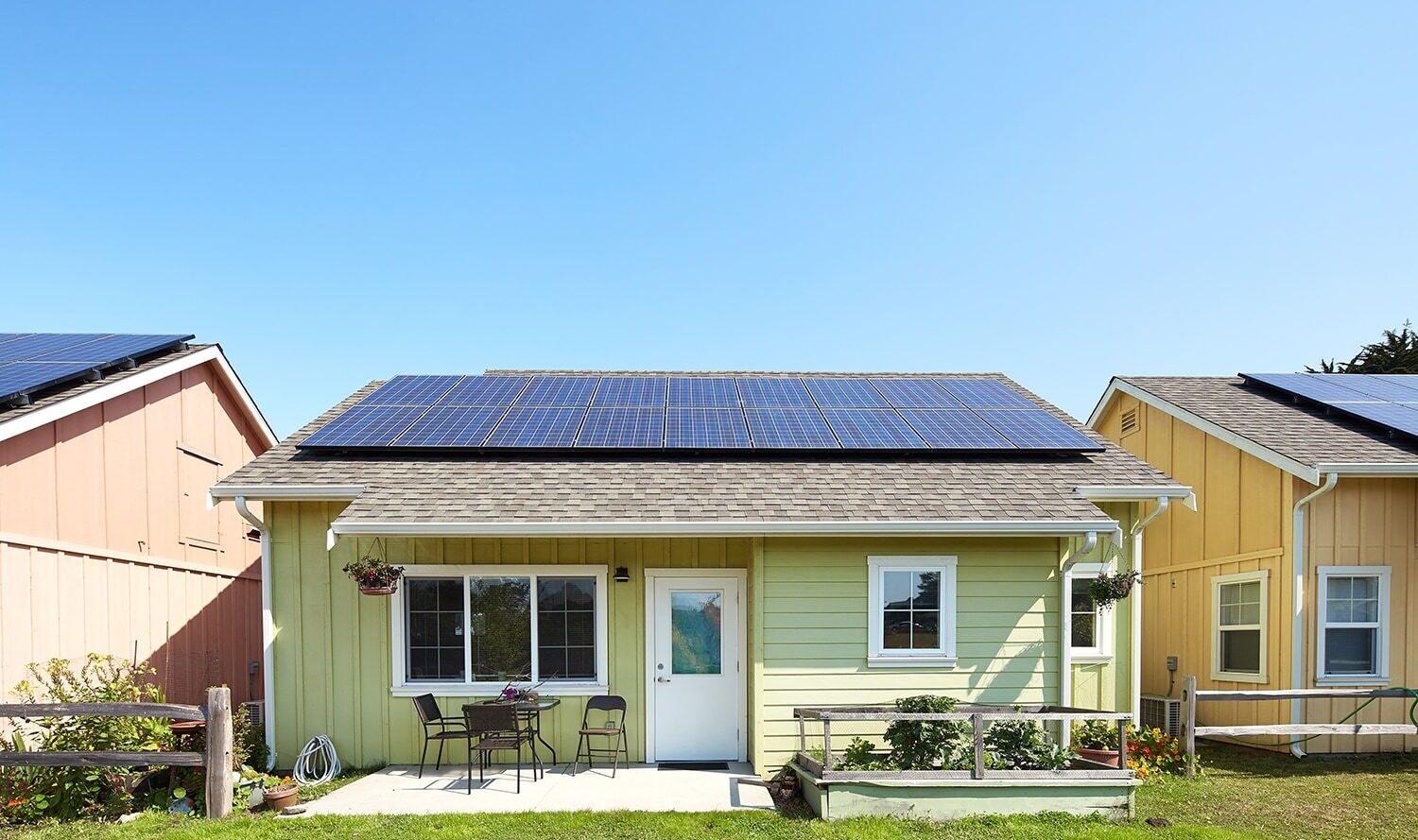 A small, single-story house with solar panels on the roof, painted light green with white trim. The front yard features a small patio set and potted plants, set against a clear blue sky.