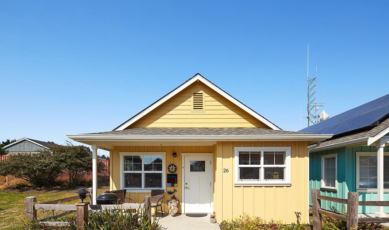 A small yellow house with a covered porch and white trim, set against a clear blue sky. There are plants and a wooden fence in the front yard, and a neighboring turquoise house is visible on the right.