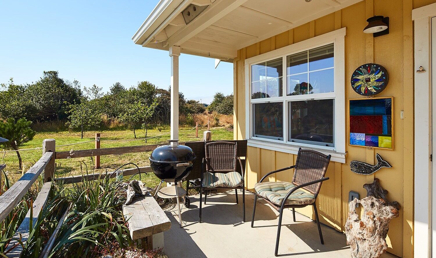 A cozy outdoor patio with two wicker chairs and pillows, a round black grill, and colorful wall art on a yellow house. The patio overlooks a green yard with trees under a clear blue sky.