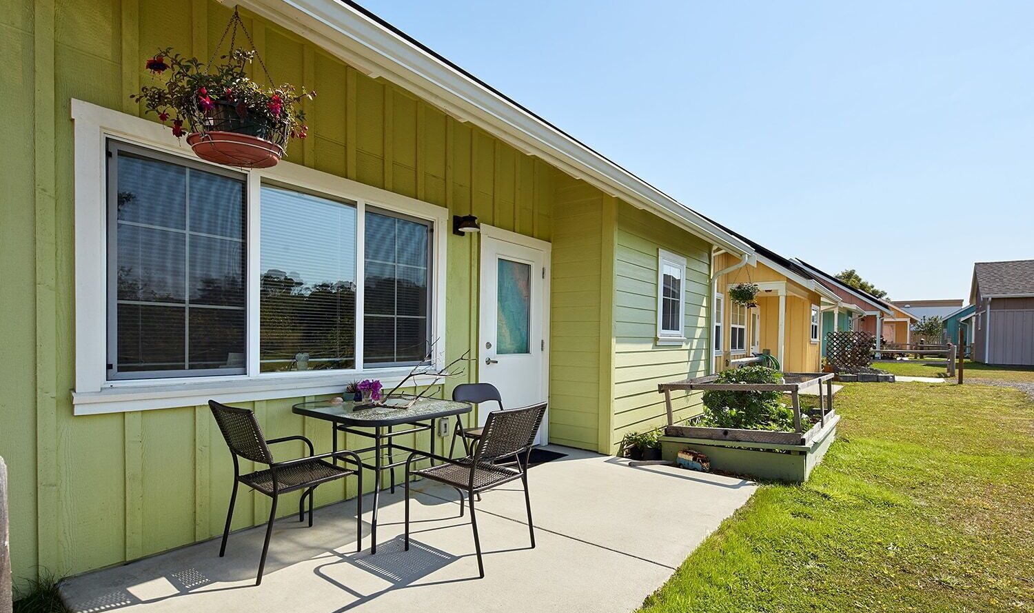 A small, single-story green house with a white door and large window. A patio with a metal table, two chairs, and a hanging flower pot is visible. The surrounding area is grassy with other similar houses in the background under a clear blue sky.
