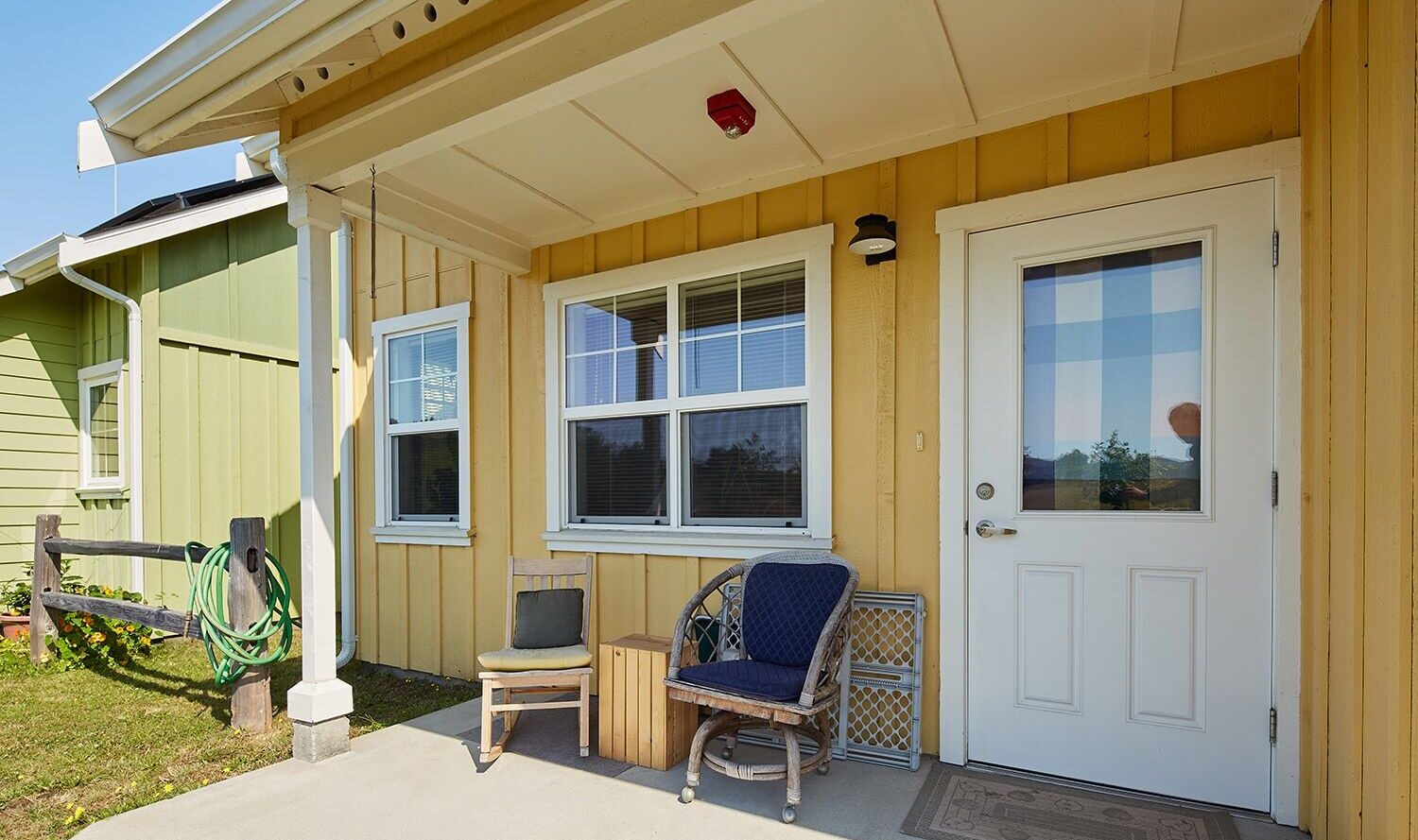 A cozy porch with a yellow wooden exterior features a white door and two windows. A wicker chair with a blue cushion and a small wooden bench sit on the concrete patio. A green hose is coiled near the house. A doormat lies in front of the door.
