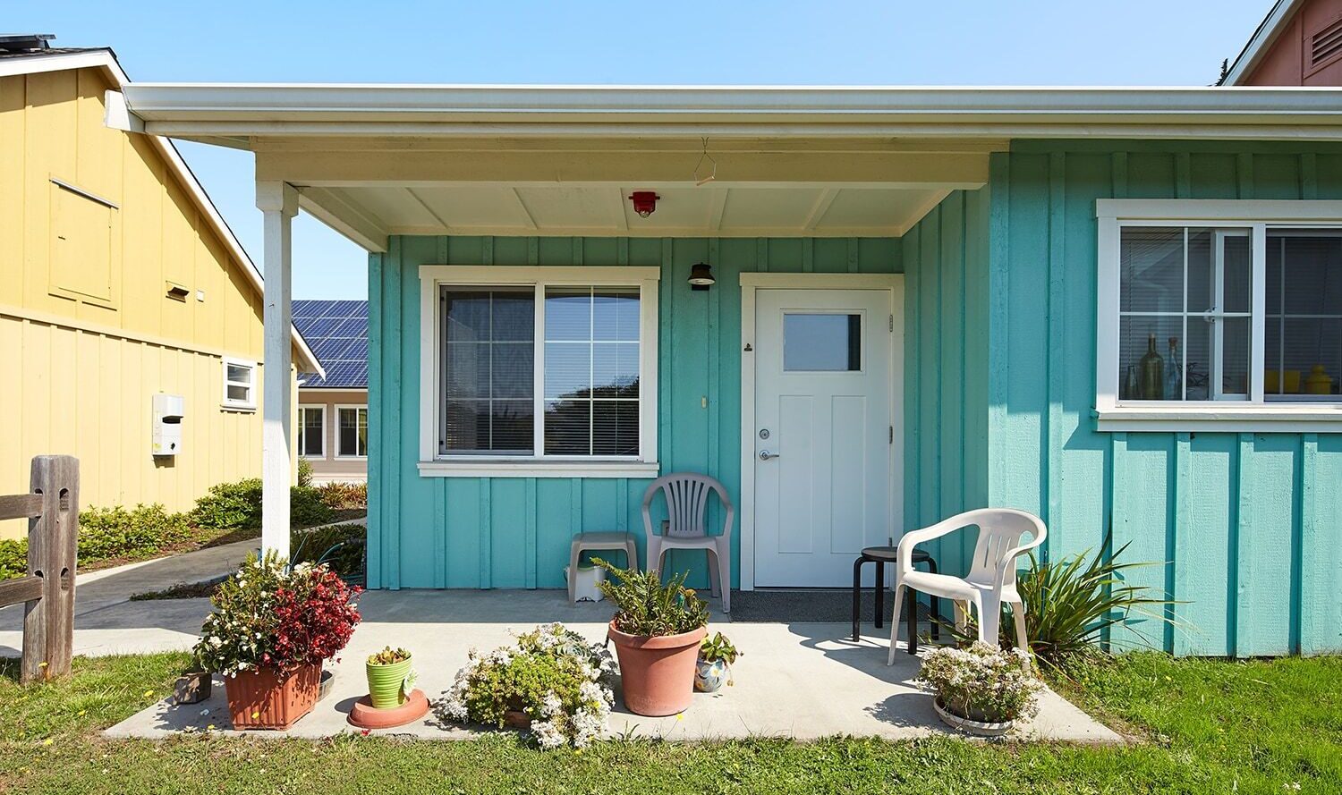 A small, light blue house with a covered porch featuring two plastic chairs. The porch is adorned with potted plants. A neighboring yellow house is partially visible on the left. The sky is clear, indicating a sunny day.