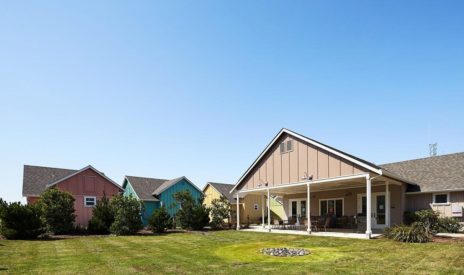 A row of colorful houses with sloped roofs stands under a clear blue sky. The houses are surrounded by manicured lawns and small bushes, with one house featuring a porch with chairs. The grass is neatly trimmed, contributing to a tidy environment.