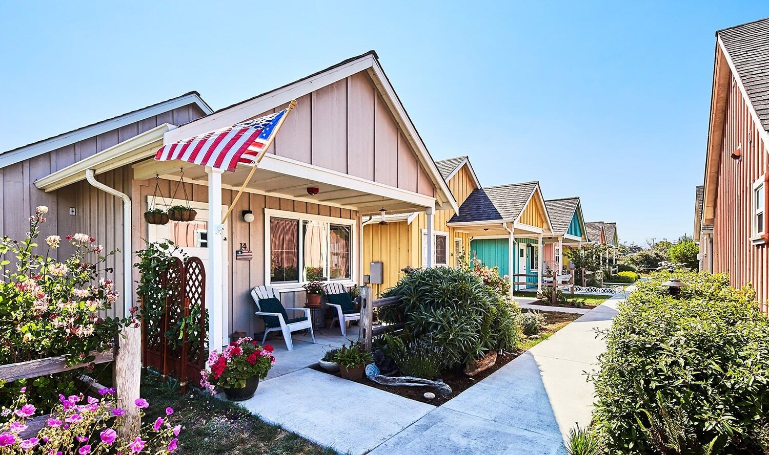 A row of small, colorful houses with porches, lined by a sidewalk and lush greenery. An American flag hangs from one porch, with chairs and potted plants adding charm. The bright blue sky enhances the vibrant atmosphere.
