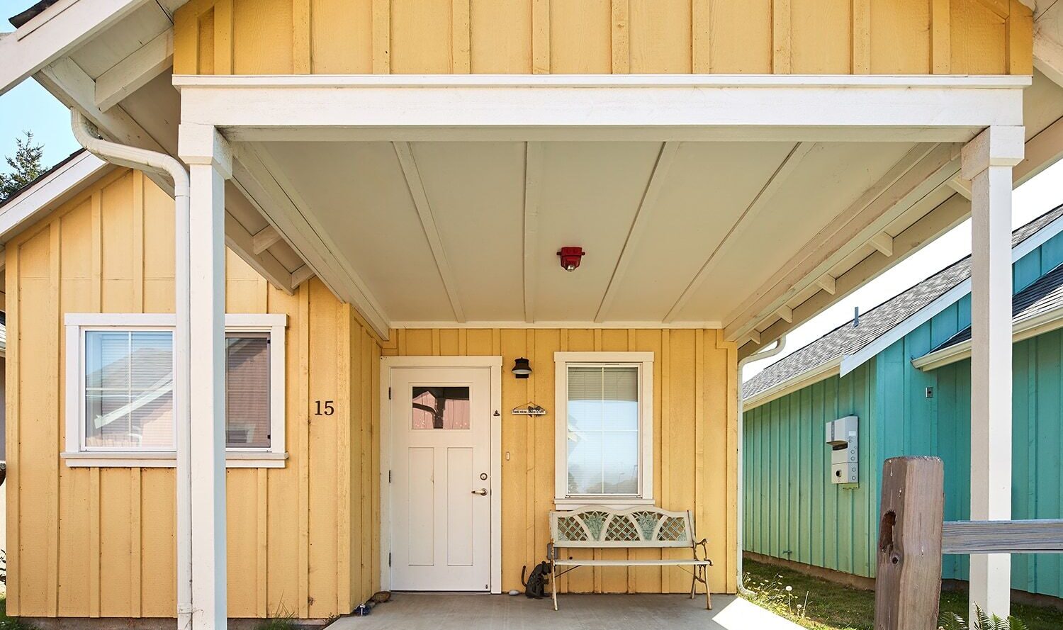 A small yellow house with a white front door, a metal bench on the porch, and a mailbox. The house is number 15, with a green house next to it. The porch is covered and theres a lawn on the side.