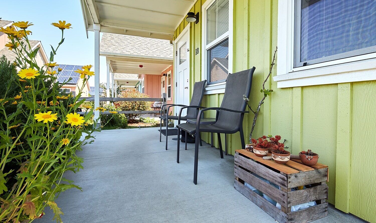 A sunny porch with two black chairs against a light green house. Yellow flowers bloom in the foreground, and a wooden crate holding clay pots is placed nearby. Other colorful houses are visible in the background.