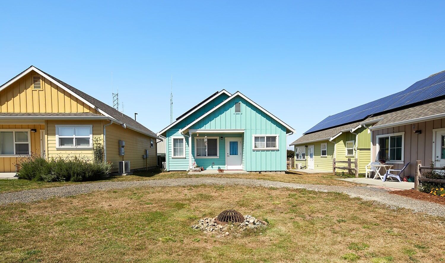 A row of small, colorful houses under a clear blue sky. The central house is turquoise, flanked by a yellow house on the left and a lighter yellow one on the right, which has solar panels. The foreground shows a grassy area with a drain.