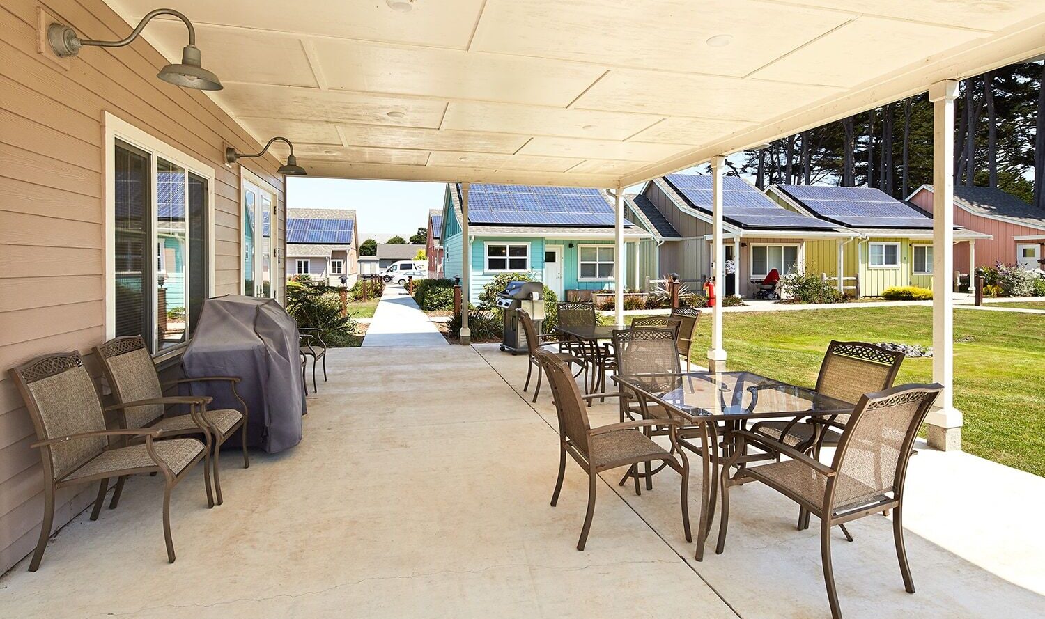 Covered patio with tables and chairs, overlooking a row of small, colorful houses with solar panels on the roofs. The area is well-lit and neatly landscaped, providing a peaceful community setting.