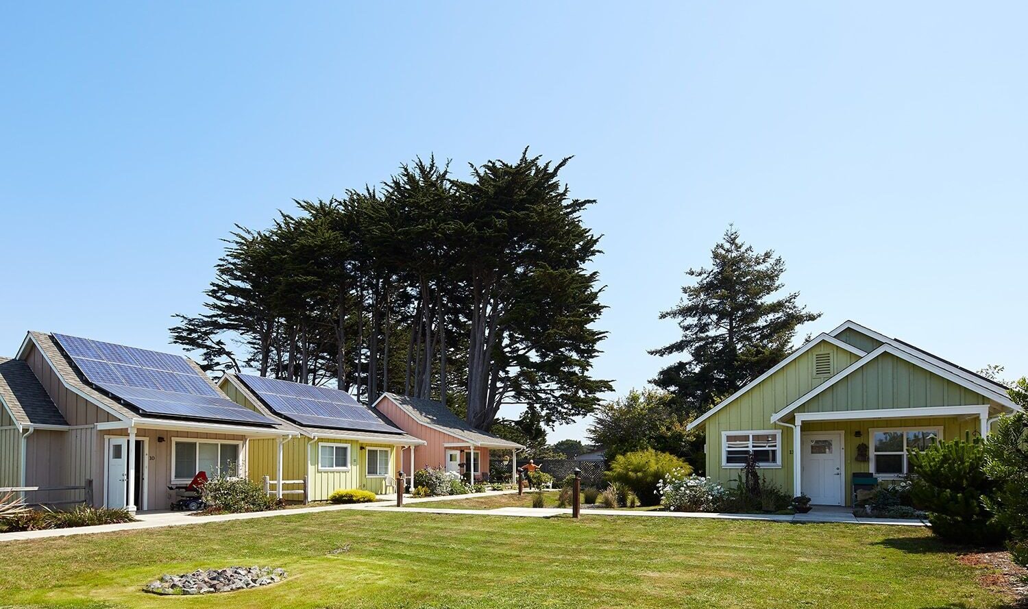 A cluster of small, colorful houses with solar panels on their roofs under a clear blue sky. The houses are surrounded by a grassy area and large trees in the background.