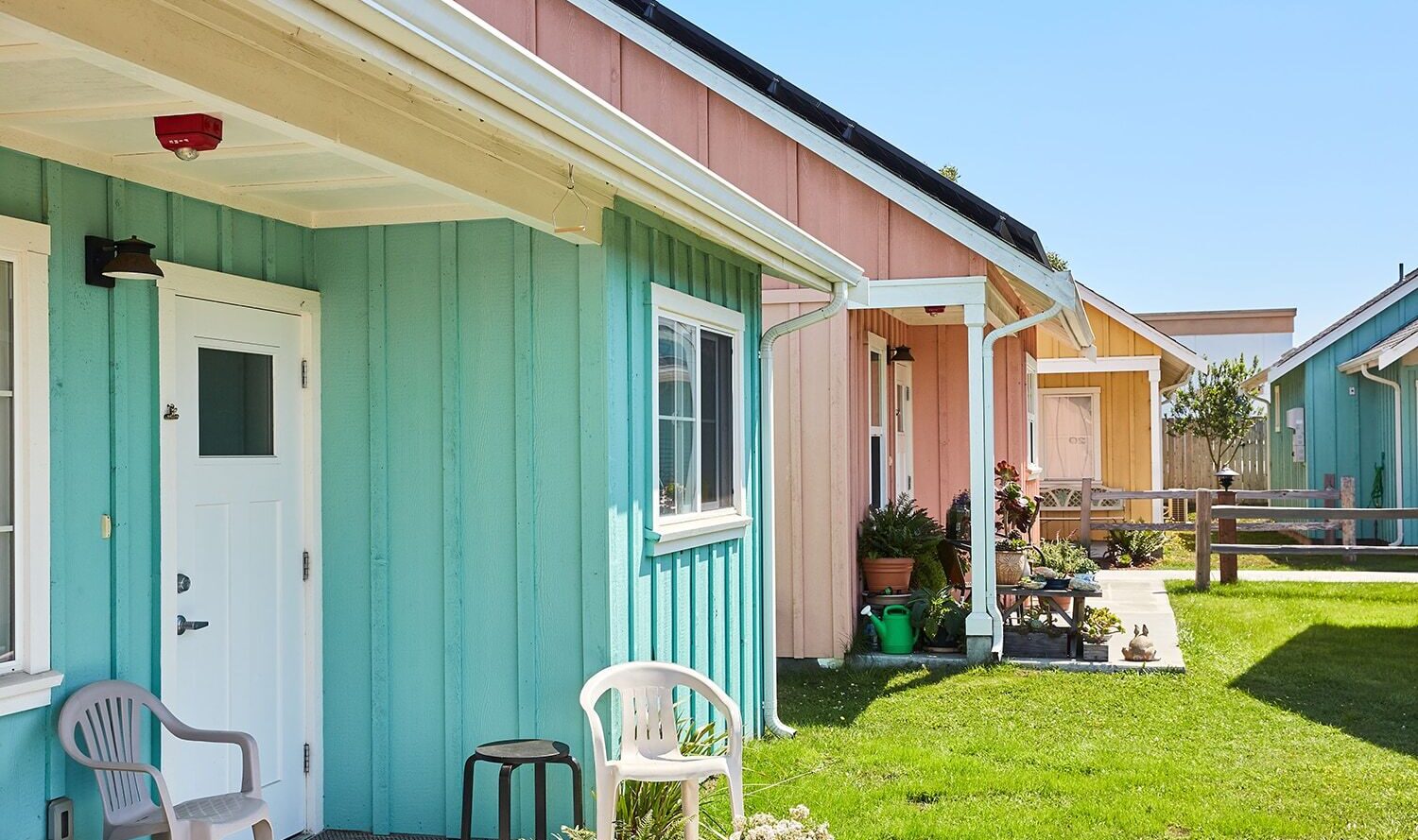 A row of colorful small houses with pastel blue, peach, and green exteriors. Each house has a small porch with chairs, and theyre surrounded by a well-maintained lawn under a clear blue sky.