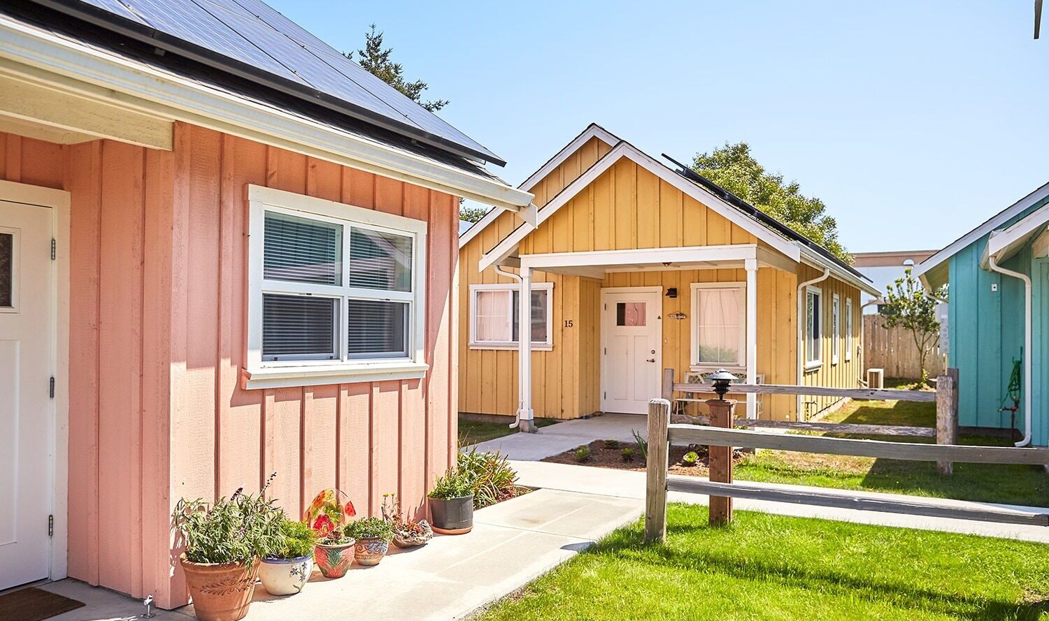 Colorful small houses with solar panels, surrounded by green grass and a clear blue sky. A pink house with potted plants in the foreground, a yellow house behind it, and a teal house on the right. A wooden fence runs along the path.