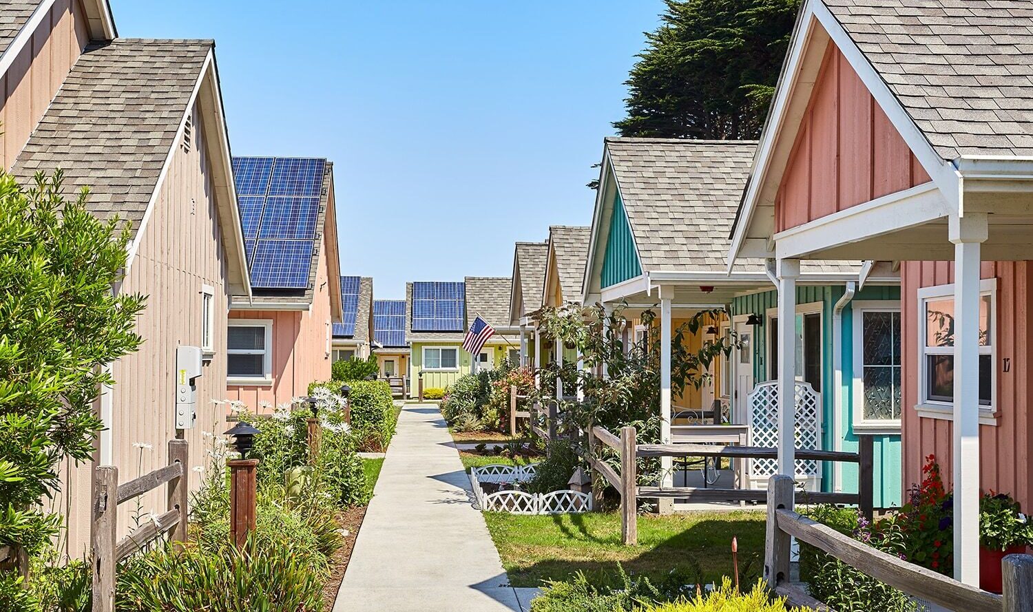 A row of colorful, quaint cottages with solar panels on their roofs line a walkway. Each house has a small garden and a picket fence. The sky is clear and blue.