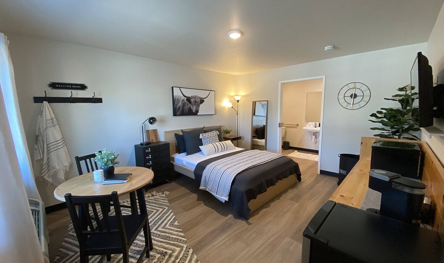 A cozy bedroom with a double bed, black and white bedding, and a wall-mounted TV. A small dining table with two chairs sits on a patterned rug. A floor lamp and potted plant add decor. Theres a bathroom visible through an open door.