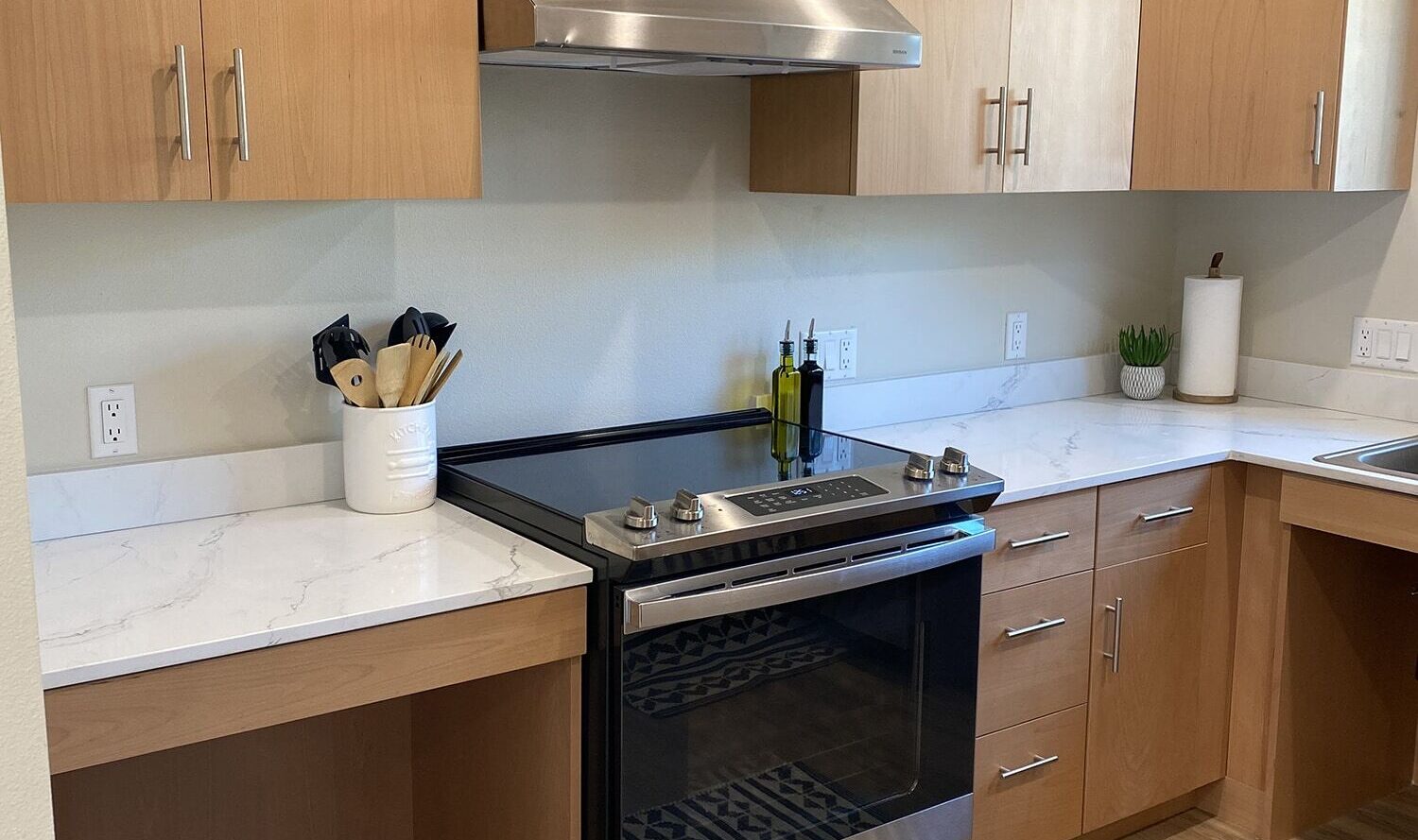 Modern kitchen featuring light wood cabinets, white marble countertops, and a stainless steel oven with a range hood. A set of utensils is placed by the stove, and a black and white patterned rug covers the wooden floor.