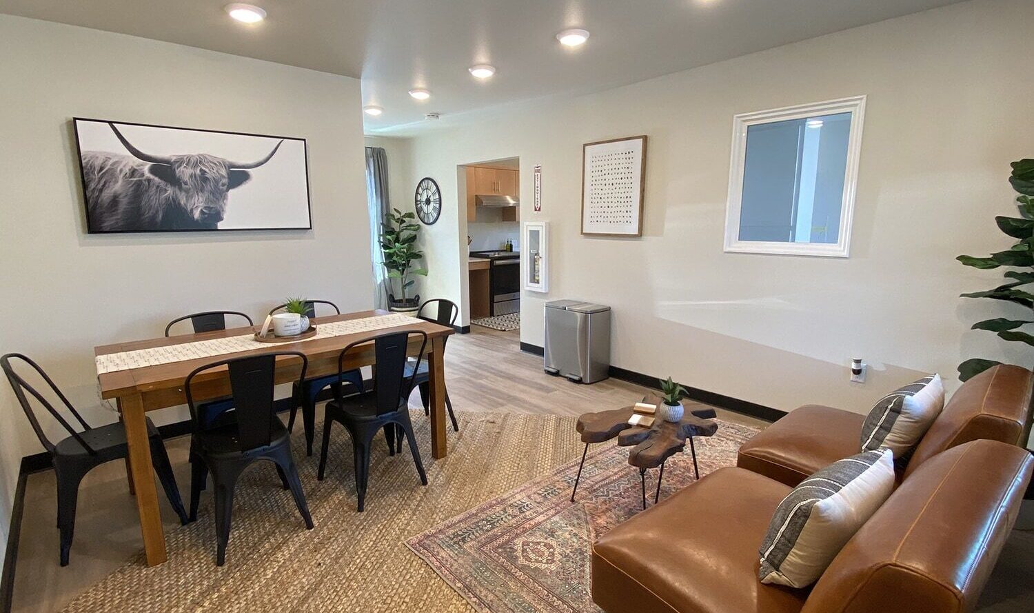 A cozy living room with a dining area. It features a wooden dining table, black chairs, and a wall art of a highland cow. Brown leather chairs, a small table, plants, and warm lighting complete the space.