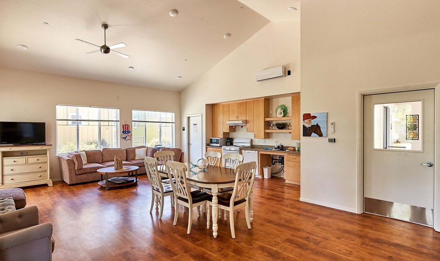 A spacious living and dining area with wooden flooring. It features a dining table with chairs, two sofas, a TV, and an open kitchen with light wood cabinets. Large windows let in natural light, and theres a ceiling fan above.
