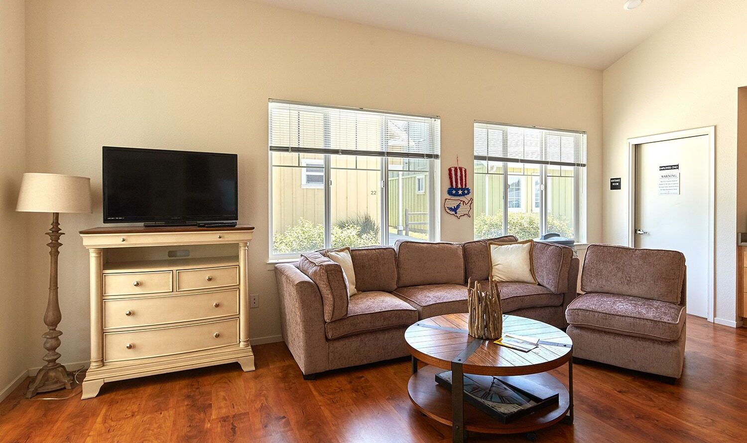 A cozy living room with a beige sectional sofa, round wooden coffee table, and a flat-screen TV on a light-colored cabinet. The room has wooden flooring and three large windows letting in natural light, along with a floor lamp beside the cabinet.