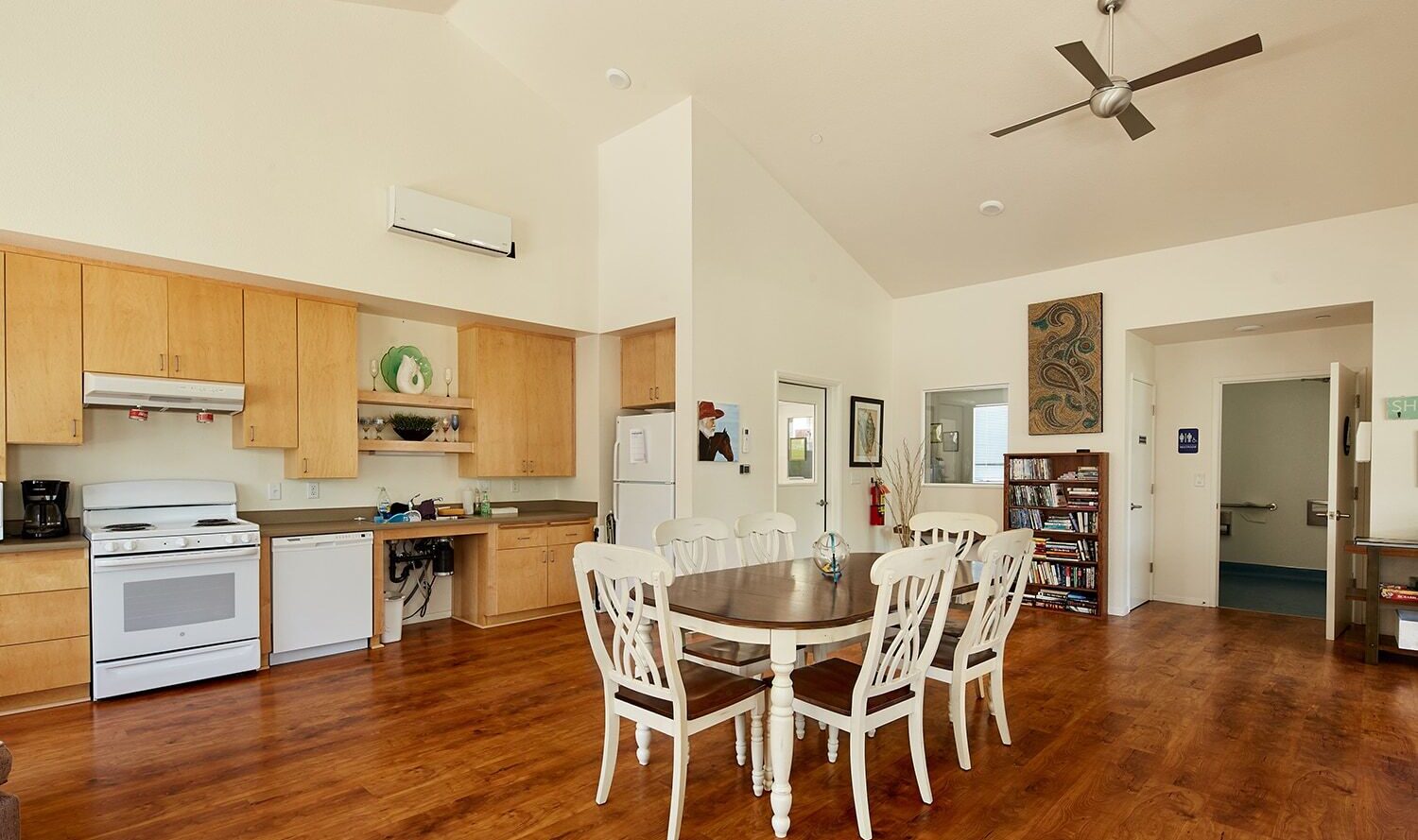 A spacious, well-lit kitchen and dining area with wooden floors. It features light wood cabinets, white appliances, a dishwasher, and a central dining table with six white chairs. Art decorates the walls, and a ceiling fan is above.