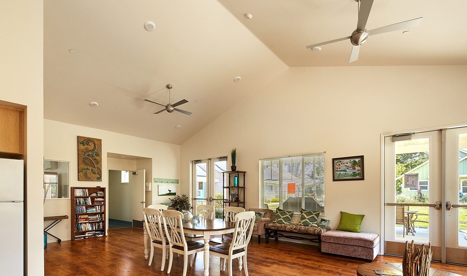 A spacious, sunlit living room with a high ceiling featuring two ceiling fans. It includes a wooden dining table with chairs, a bookcase, a cushioned bench by the window, and a glass door leading outside. Walls are adorned with artwork and decor.
