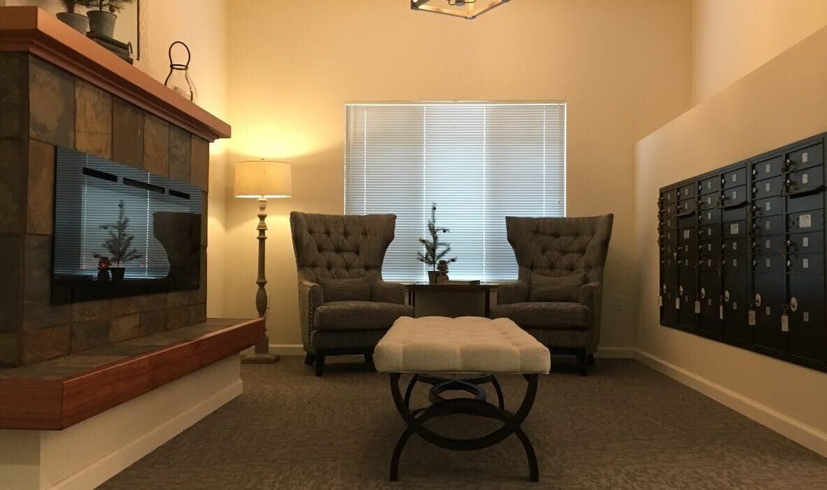 A cozy room with two tufted chairs and a table in front of a window, a stone fireplace on the left, and a row of black mailboxes on the right. A cushioned bench is centered on the carpeted floor beneath a soft light.