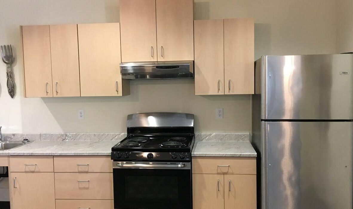 A kitchen with light wood cabinets, a stainless steel fridge, oven, and range hood. A marble countertop houses a sink on the left. Two decorative utensils hang on the wall.