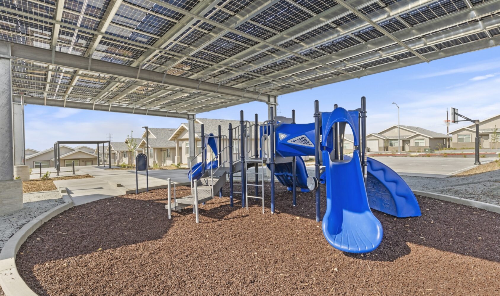 A playground with blue slides and climbing structures on wood chips, beneath a canopy of solar panels. Nearby are houses and an empty basketball court, all under a clear blue sky.