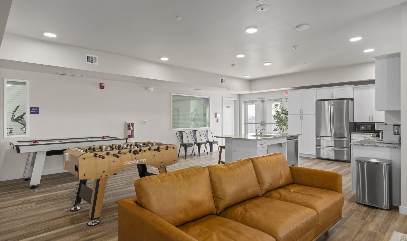 A modern game room and kitchen area featuring a brown sofa, foosball table, air hockey table, and high-backed chairs at a dining counter. The kitchen includes stainless steel appliances and white cabinetry. The room has wooden flooring and bright lighting.
