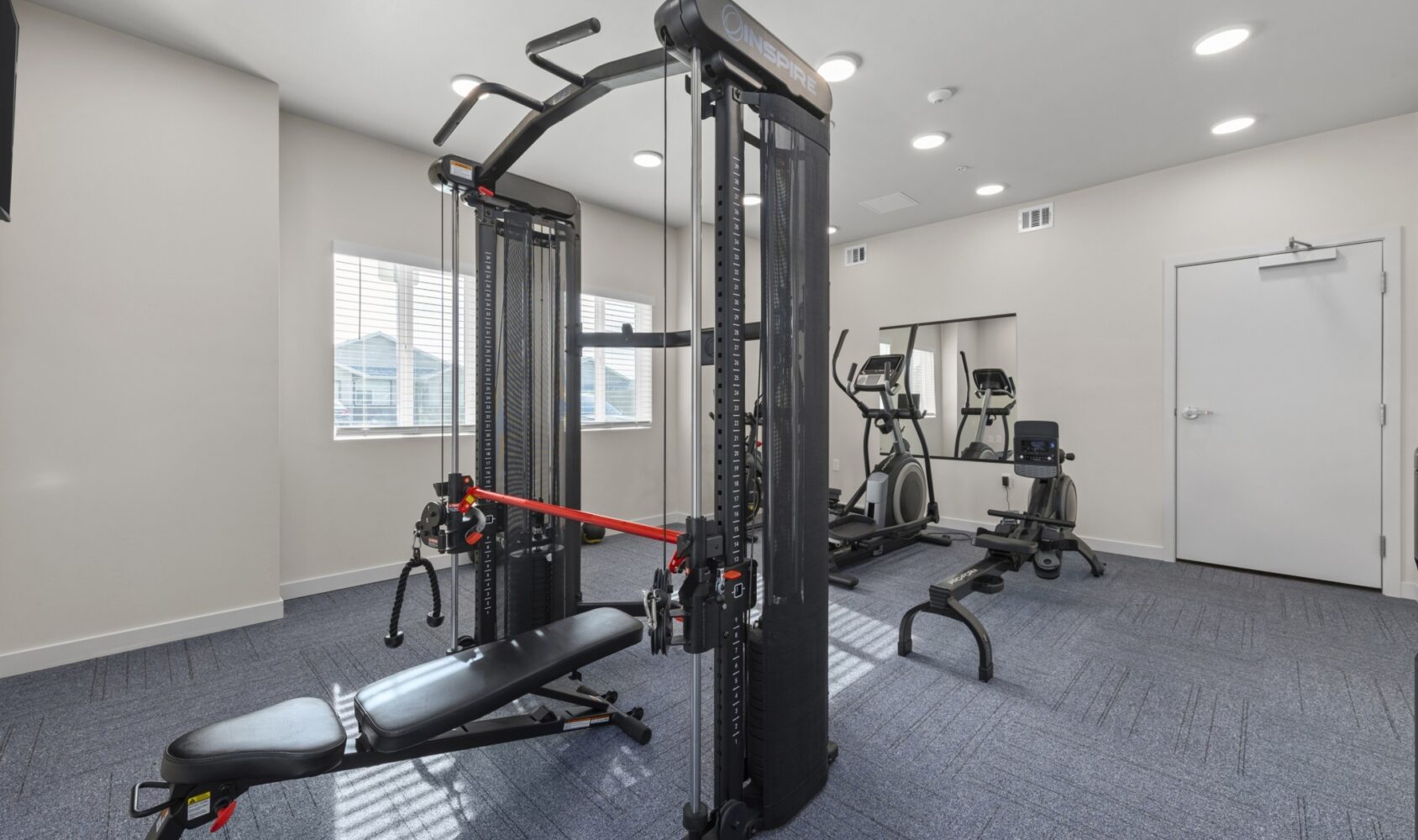 A gym with a weight machine, exercise bike, and elliptical trainer. The space is well-lit with natural light from a window and features blue carpet and a large mirror on one wall.