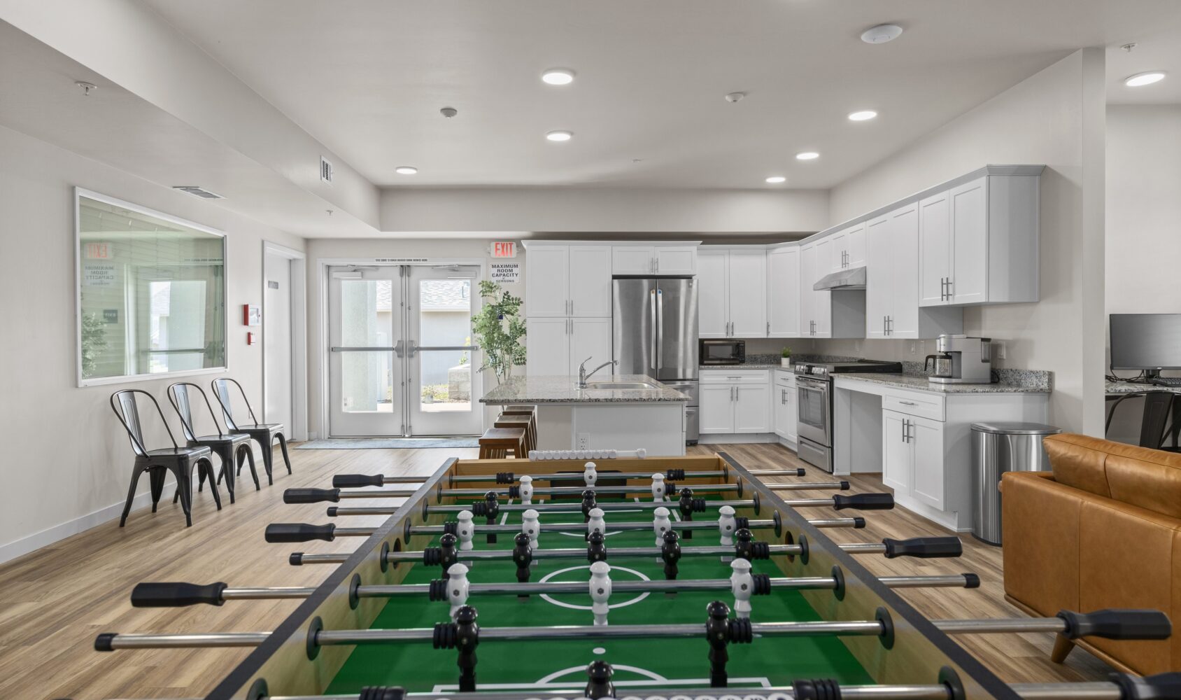 A modern recreation room with a foosball table in the center. The room also features a kitchen area with white cabinets, stainless steel appliances, and a seating area with metal chairs. Large windows provide natural light.