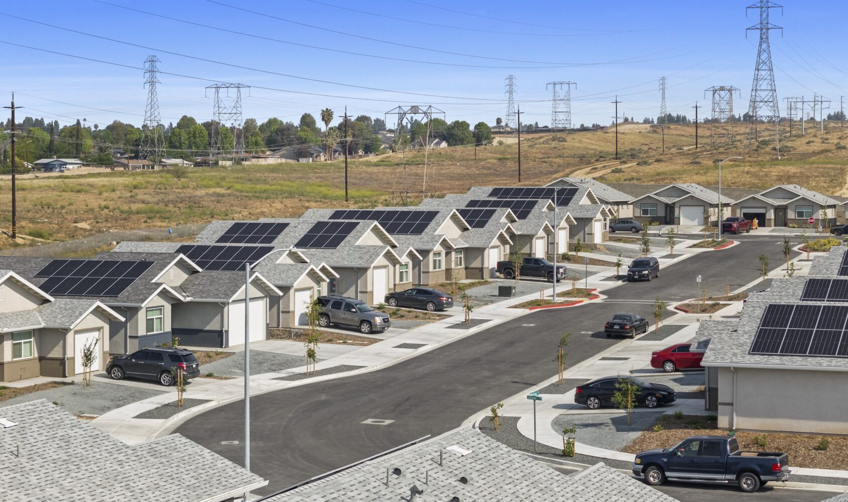 A suburban neighborhood with single-story homes, each equipped with solar panels on the roofs. Several cars are parked in driveways along a winding road. The area is surrounded by open fields and electricity pylons in the background.