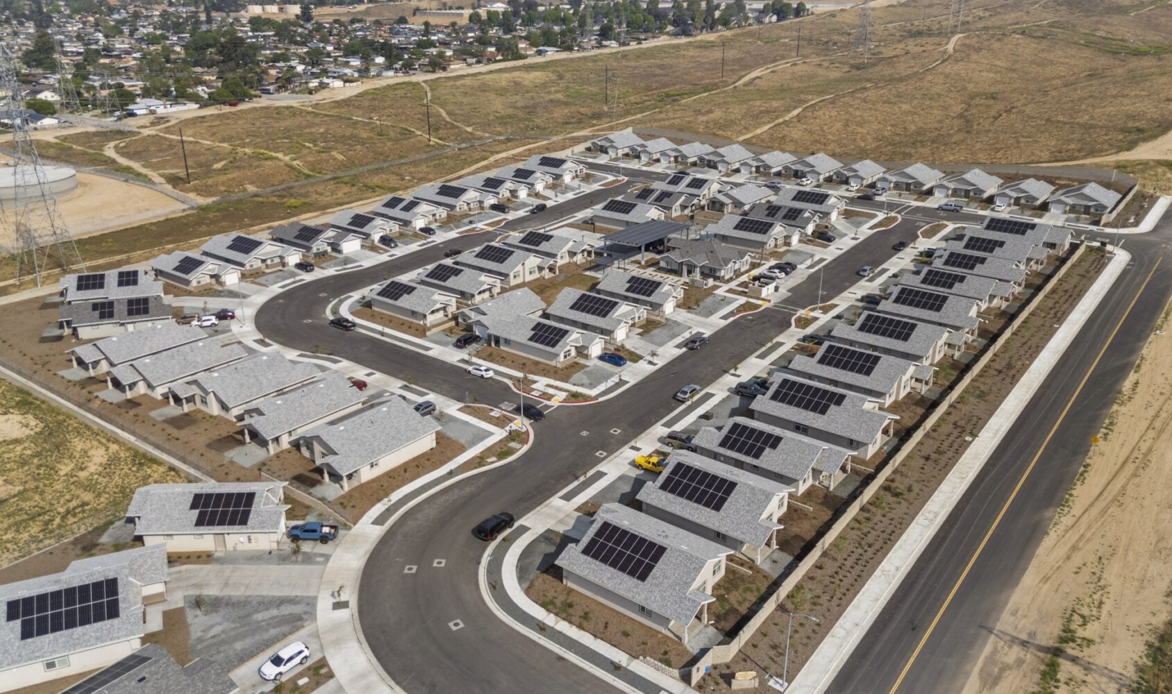 Aerial view of a suburban neighborhood with rows of identical white houses, each equipped with solar panels on the rooftops. The area is surrounded by undeveloped land and a network of paved roads.