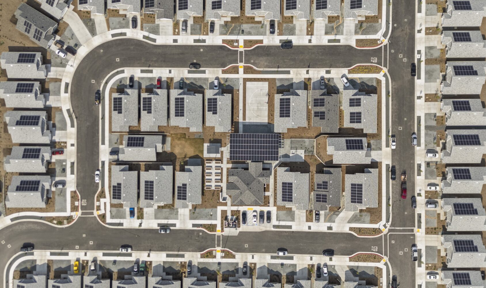 Aerial view of a suburban neighborhood with rows of houses, all featuring rooftop solar panels. The houses are arranged along curving roads with driveways, and small yards surround each property. The streets form a grid-like pattern.