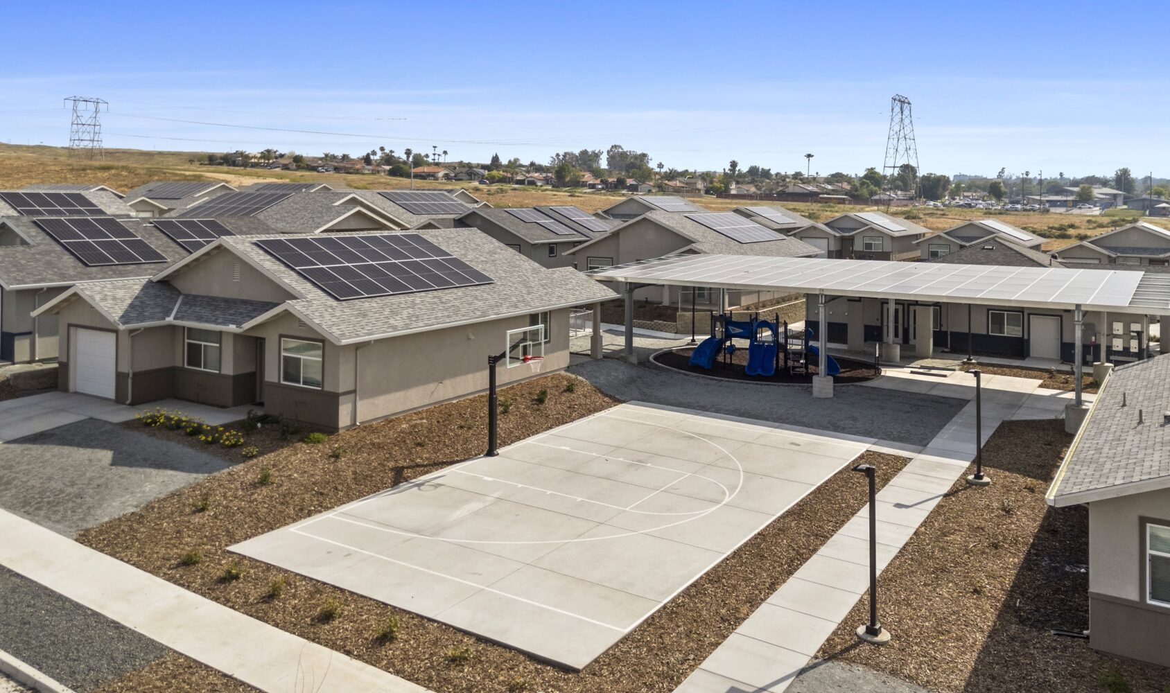A residential area with houses featuring solar panels on roofs. A basketball court and a covered playground area are visible in the center. The landscape is dry with scattered houses, and power lines can be seen in the background under a clear blue sky.