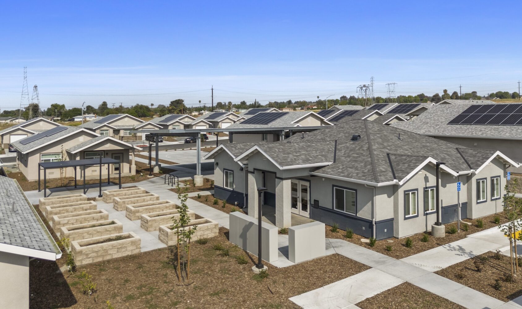 A residential complex featuring modern single-story homes with gray roofs and light-colored exteriors. Each unit has a small yard and a paved walkway. Solar panels are visible on some roofs, and there is a view of a landscaped communal area.