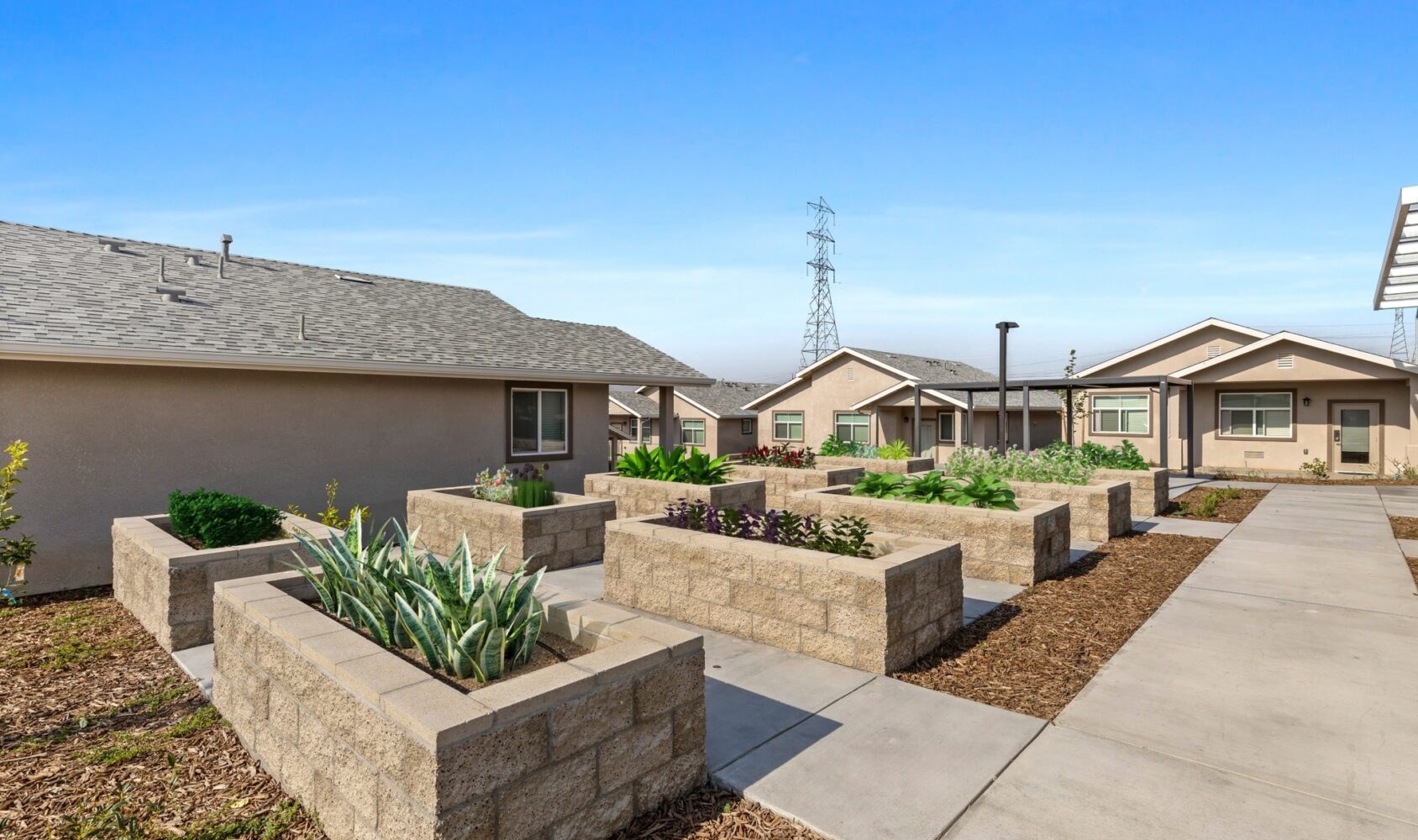 Community garden with raised stone planters filled with green plants. The area is surrounded by single-story houses under a clear blue sky. Paved walkways connect the spaces, and an electrical tower is visible in the background.